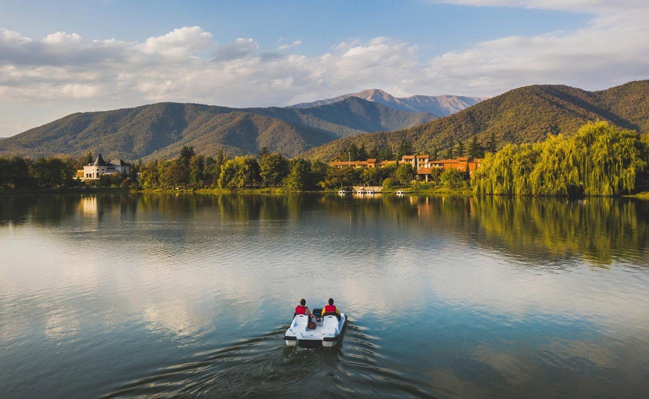 Two people on a pedal boat in a lake surrounded by mountains and trees under a cloudy sky.