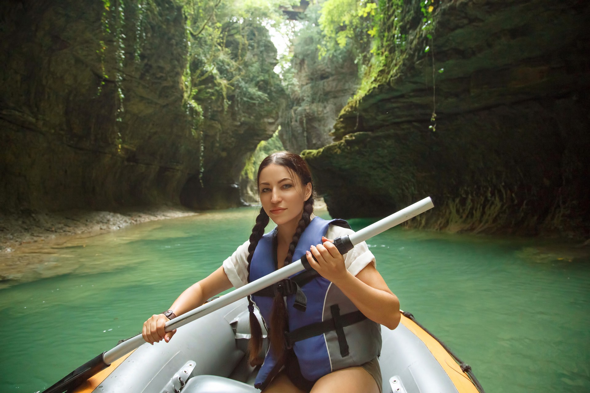 pretty young woman with dark hair gathered in two black braids, lady looks and poses for camera in life jacket and paddle in hands, tourist in inflatable boat sails through Martvili Kanyon Georgia